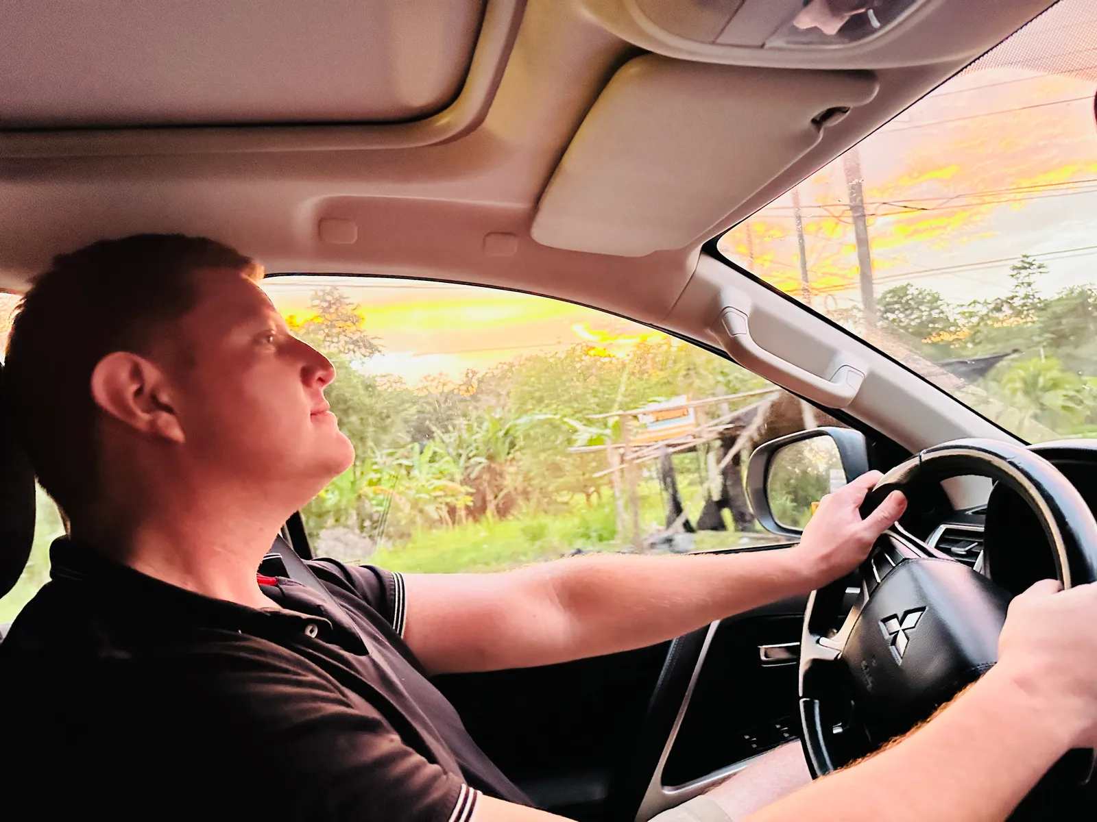 A man at the wheel of a car seen from the passenger seat, with a sunset sky outside.